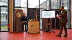 Moderator Andreas Kaufmann steht im Saal vor dem Informationsstand der Agil und spricht zu einer Vertreterin und einem Vertreter der Agil.