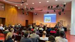 Ein Mann am Rednerpult auf der Bühne spricht zum Publikum im Saal, dass auf Stühlen in Reihen sitzt.