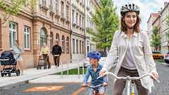 Kommunaler Wäremplan Stadtumbau Header Visualisierung: Ein Frau fährt mit einem Kind Fahrrad auf einer Straße. Auf dem Gehweg eine älter Frau mit Rollator und eine Mutter mit Kinderwagen.