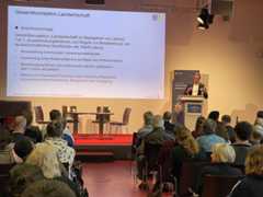 Sylvio Etling steht am Rednerpult auf der Bühne und spricht zum Publikum. Im Hintergrund ist seine Präsentation zum Thema Landwirtschaftskonzept zu sehen.