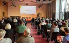 Ein Mann am Rednerpult auf der Bühne spricht zum Publikum im Saal, dass auf Stühlen in Reihen sitzt.