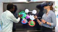 Mann in weißem Hemd und Frau mit 3D-Brille und Eingabegeräten vor großem Computerbildschirm, der eine 3D-Grafik aus bunten. molekülartig verbundenen Kugeln zeigt.