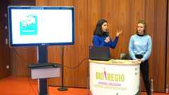 Zwei Mitarbeiterinnen der Bio-Regio-Modellregion Westsachsen stehen an ihrem Infostand. Eine von Ihnen spricht in ein Mikrofon. Links neben Ihr steht ein Präsentationsmonitor auf einem Rollgestell.