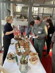 Eine Frau steht an einer weiß gedeckten Tafel mit Brot und Olivenöl und spricht zu einem Mann.