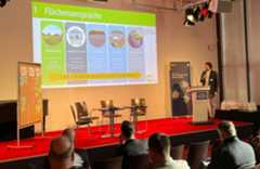 Ein Mann steht auf einer Bühne und spricht ins Mikrofon. Vor der Bühne sitzt das Publikum und hört zu.