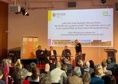 Eine Frau steht auf der Bühne und spricht zu drei Männern, die neben ihr sitzen. Im Saal davor sitzt das Publikum und hört zu.