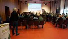 Hinter den Sitzreihen mit Publikum steht ein Teilnehmer mit Mikrofon in der Hand und spricht die drei Gesprächsteilnehmer auf der Bühne an.