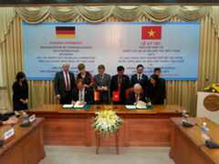 In einem Raum mit Bankettbestuhlung steht eine Gruppe von Damen und Herren an einem Tisch hinter den beiden Herren, die ein Papier unterzeichnen. Im Hintergrund sind beschriftete, blaue Tafeln mit den Flaggen von Deutschland und Vietnam an einem grünlichen Vorhang befestigt.