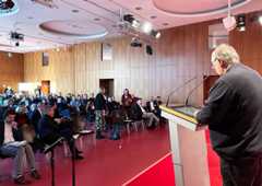 Ein Mann am Rednerpult spricht von der Bühne zum Publikum im Saal.