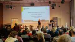 Die Moderatorin mit Mikrofon in der Hand steht auf der Bühne und spricht zu einem Mann am Rednerpult.