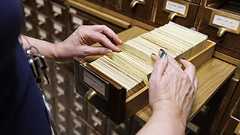 A person reading a book kneeling in front of a card catalog.
