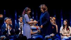 Eleonora Svanberg in a lavender dress with a floral hair accessory shakes hands with Charlotte Wall, Vice President of the Anders Wall foundation in a navy blue suit on a stage. Eleonora is smiling. Behind them, a group of people, dressed in formal attire, are seated and clapping. The men are wearing suits with red flower pins, while the women are in elegant dresses, some also adorned with red flowers. The stage is lit with blue lighting and spotlights, giving the scene a formal and celebratory atmosphere.
