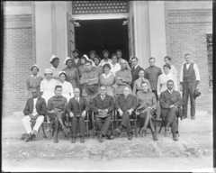 A black and white photo of a group of people sitting and standing in front of a building entrance wearing formal and nursing attire