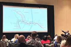 A speaker at a podium gestures at a large projection screen showing the MetroLink system map during a presentation to a group