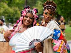 Close-up of two beautiful Black women posing wearing colorful clothing. One holds a large white folding fan.