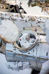 An astronaut in a white spacesuit comes out of a hatch on the International Space Station. We can see parts of the space station in the photo.