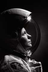 A black and white photo of a woman wearing a spacesuit and helmet. She looks upward toward the right. The photo shows only her head and shoulders.