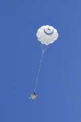 A parachute carrying a capsule is fully open against a canvas of deep blue sky.