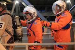 NASA astronauts Christina Koch, a white woman, and Victor Glover, an African-American man, walk to the left during a night-run launch day demonstration. The crew access arm they're walking on has railings, one of which is in the foreground. The astronauts are wearing orange suits with patches and straps on them, as well as white helmets with clear visors. There is a person walking in front of them. This person is wearing khaki clothing and is carrying a gas tank on their back. The gas tank has straps like a backpack.