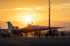 NASA’s X-59 quiet supersonic research aircraft is seen on the ramp at sunrise with its canopy open. Orange and gold light filters through the clouds behind the aircraft, highlighting the aircraft’s shape. Several ground crew members are visible around the aircraft, along with support carts. A vertical light pole with a coiled cable can be seen in the foreground.