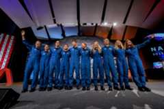 Ten people in blue jumpsuits link arms. The two women on either end wave at the audience, who is not visible.