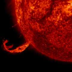 This image focuses on a twisting jet of material erupting from the Sun, glowing bright against the black backdrop of space. The Sun is a fiery, deep red ball taking up three-quarters of the image on the right.