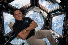 A man in a dark polo shirt smiles at the camera with his arms crossed. He is in the cupola of the International Space Station, an area with multiple windows through which Earth and space can be seen. Earth's clouds can be seen through the windows behind him.