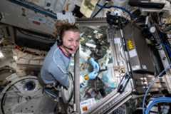 An astronaut smiles at the camera while she works at a glovebox on the International Space Station. Her hands are inside the glove box. She is wearing a black headset.