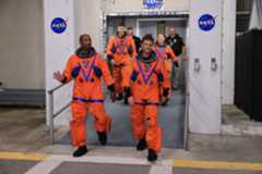 Four astronauts - three men and one woman - walk down a dark gray ramp, exiting a building. A Black man at front left waves as he looks to our right. The other three people are smiling. All of the astronauts wear bright orange jumpsuits with various patches on them.