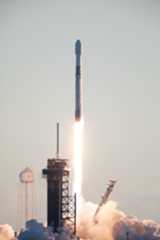 A rocket lifts off into the sky, leaving a bright trail of flame in its wake. White vapor clouds billow out along the ground. There are also various structures in the image, including a gantry and a white structure that looks like a water tower.