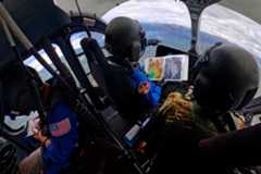NASA astronauts Matthew Dominick (left, in a blue jumpsuit with a patch on the right arm) and Mark Vande Hei (right, in camouflage clothing) in the cockpit of a helicopter. Dominick holds an open binder in his right hand and points forward with his left. They are both wearing black helmets. There is another person behind Dominick, also wearing a blue jumpsuit and helmet, facing the opposite way.