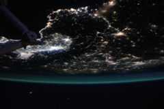 Earth is a dark sphere with a glowing green border in this photo. We can only see one portion of it; in this photo’s orientation, we’re looking at the “bottom” of Earth’s sphere. City lights shine brightly at middle left; they are more dispersed throughout the rest of Earth that’s visible. Part of the space station is just barely visible at top left.