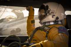 A person facing left sits inside a vehicle. They are wearing a yellow jumpsuit and gloves and a white helmet with a visor that looks like a smaller version of the kind astronauts wear during spacewalks. Outside of the window behind the person, you can see the ER-2 aircraft being prepared for flight.