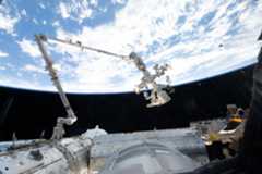 The 57.7-foot-long Canadarm2 robotic arm extends from the International Space Station. The arm is long, cylindrical, and white, with several joints that are bent to the right (at left in the image) and downward (at center). Earth makes a bright white and blue background for the top half of the photo. Our home planet contrasts starkly with the darkness of space.