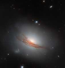 A disk-shaped galaxy. It glows brightly at the center and shines a faint white light all around it. The disk is made up of tightly-packed rings of dust, some darker and some lighter. Wide, long lanes of dark reddish dust cross the galaxy in front of its edge, blocking out some of its light; the long strands twist and break apart at each side. A couple of nearby stars and distant galaxies are also visible on the black background.