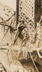 A man stands on a plank, which is attached to the bridge by ropes. He is holding a rivet gun against the metalwork, which is covered in rivets. - click to view larger image