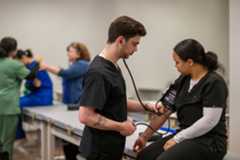 Two medical students in scrubs. One is using a stethoscope to test the others vitals