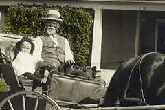 An older man in a waistcoast, pants, shirt, and a hat sits next to small white child in a horse-drawn buggy.