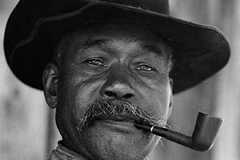 Portrait of Adam Grayson wearing overalls, a dark hat, and smoking a pipe.