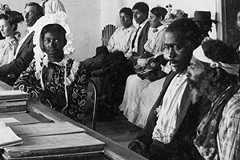 Several men and women sit in a large room behind a wooden bar. A few individuals sit at large desks on the other side of the bar.