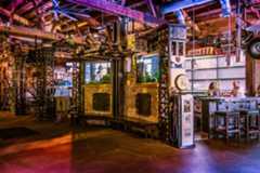 Interior of a themed bar with Route 66 decor, vintage car parts, and industrial elements. Two people sit at a metallic bar with stools, under exposed wood and metal beams.