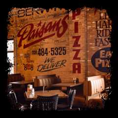 Restaurant interior with brick wall featuring large painted signage for Paisans Pizza, including phone number and "We Deliver" text. Booth seating with tables in foreground.