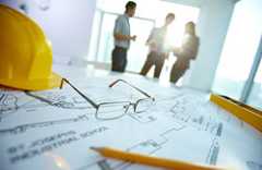 Mesa com papeis, capacete de obras amarelo e lápis. Ao fundo 3 pessoas conversam.