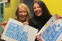 Two women holding signs thanking people for signing up and supporting Dry July, in front of a bright yellow wall.