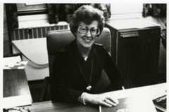 A woman in glasses, Loretta Ford, sits at a desk surrounded by papers, reflecting on her time as a former nursing dean.