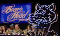 Close up of an ice sculpture of a boar's head with a screen in the background that reads "Boar's Head Dinner" and faculty in medieval garb at a high table.