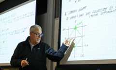 David Foster in a classroom teaching and gesturing at a screen full of graphs and equations.