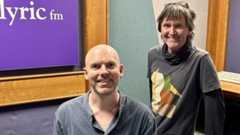 Composer Michael Gallen sitting in a radio studio and presenter Paul Herriott standing beside him, both smiling.