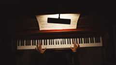 Birds-eye view of a person playing an upright piano in a dark room. A music book is open in front of them.