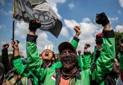 Outside, beneath a blue sky with fluffy white clouds, people wearing bright green jackets hold their fists in the air and wave a flag.